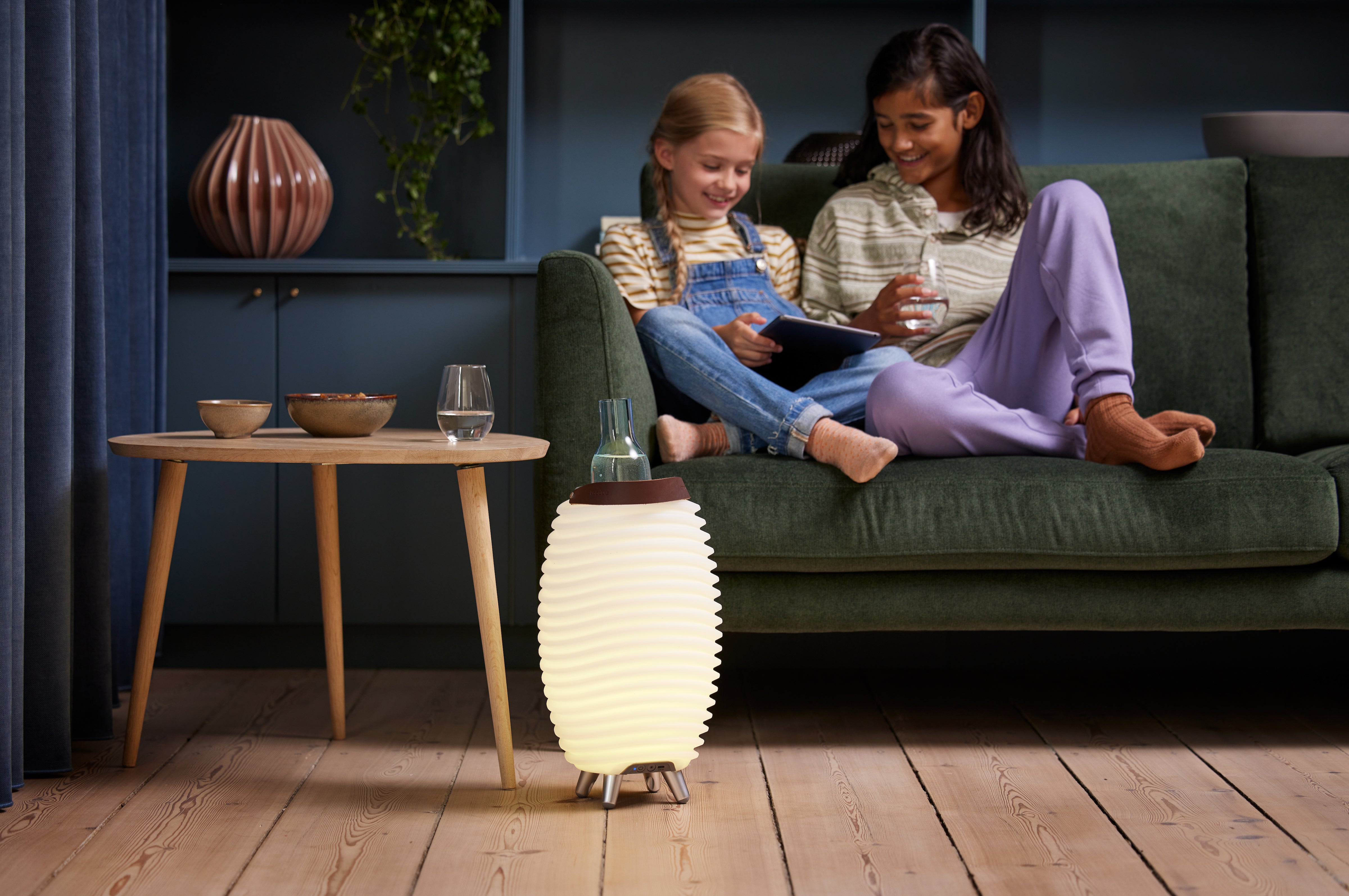 synergi lampe plassert med en flaske i kjøleren, på et gulv foran en sofa med 2 jenter sittende