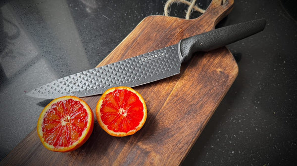 kniv med sort skaft fra royalty line liggende på en brun skjærefjøl med en delt blodappelsin ved siden av kniven