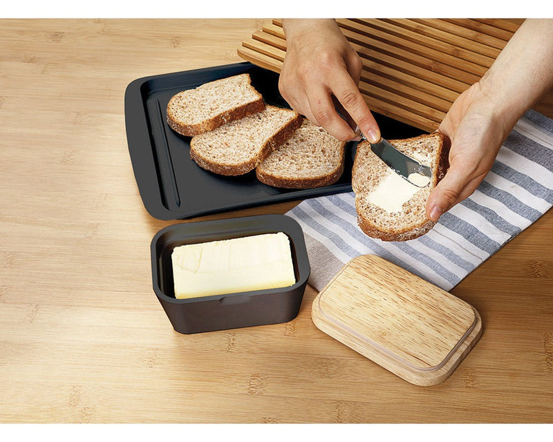 2 hender smører en skive brød over et bord hvor smørboks, lokk og et fat med brøskiver er plassert på en blåstripete duk