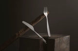 brimi kniv og gaffel i stål på en trestubbe lent mot en grein