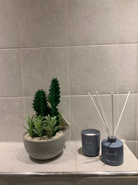 duftpinne og duftlys på en baderomshylle ved siden av en kunstig plante i grå potte