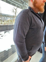 mann med skjegg ikledd en merinoulljakke i blå farge foran et vindu med snø utenfor på terassen