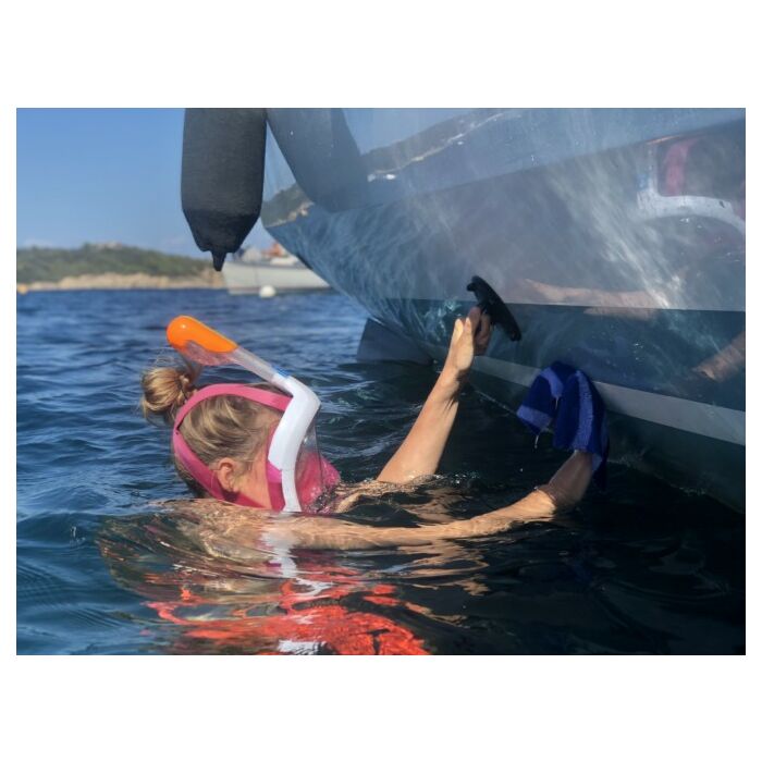 kvinne med dykkemaske og snorkel i vannet som holder seg festet til en båtside samtidig som hun vasker båten med høyre ledige hånd
